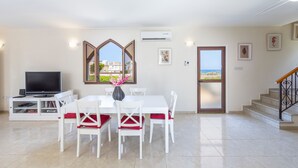 A modern living room with a dining table, white chairs, a flat-screen TV, and a large window with a view of the sea. - Protaras Villa Elena 3 (Protaras)