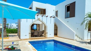 A swimming pool with a blue interior, surrounded by a white building with a covered patio area. - Protaras Villa Elena 3 (Protaras)