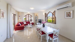 A modern living room with a red sofa, white dining table, and chairs, a TV, and a bookshelf. - Protaras Villa Elena 3 (Protaras)