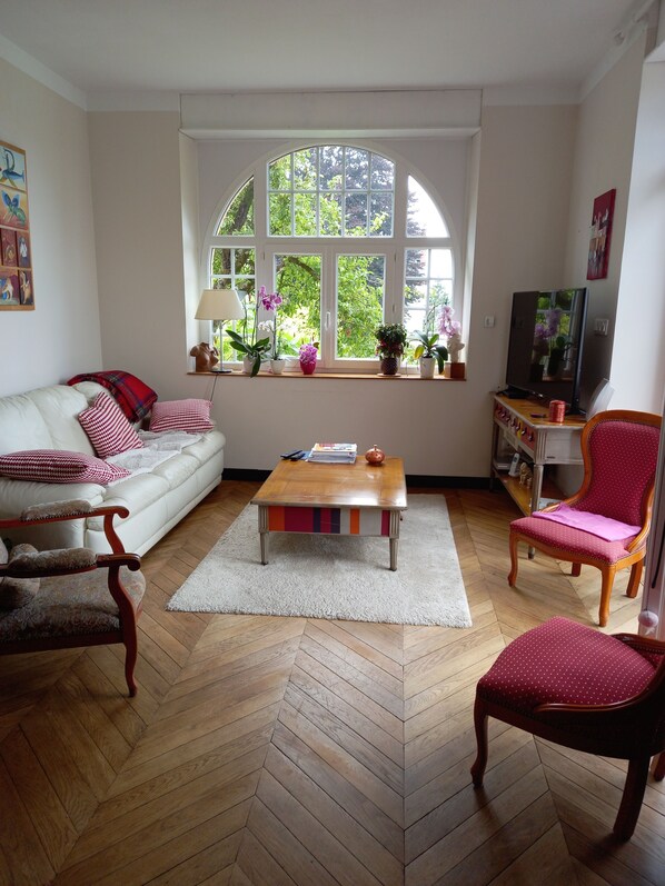 Ein Wohnzimmer mit einer Couch, Sesseln, einem Couchtisch, einem Fernseher und einem großen Fenster mit Pflanzen.