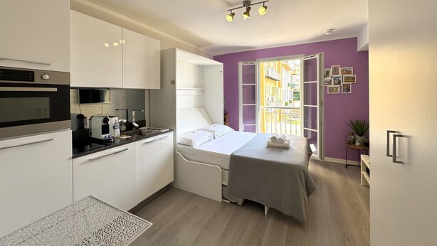 A compact hotel room with a kitchenette, a bed with a grey bedspread, a window with a view of buildings, and a small potted plant. - Maison Linda (Niza)