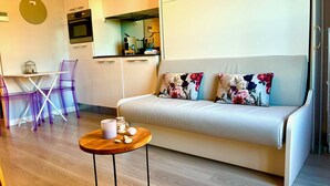 A modern living room with a sofa, a small wooden table with a mug and cookies, a purple chair, and a kitchen area in the background. - Maison Linda (Niza)