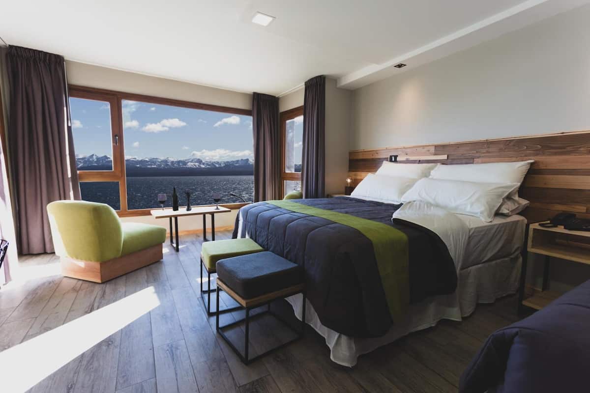Ein modernes Hotelzimmer mit großem Fenster und Blick auf die Berge, einem Bett mit Holz Kopfteil, einem grünen Sessel und einem kleinen Tisch.