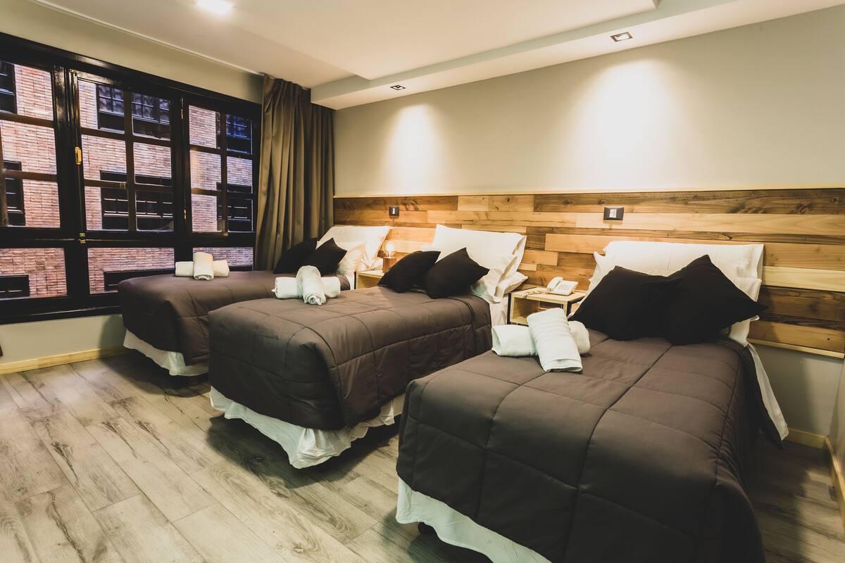 Ein Hotelzimmer mit zwei Betten, einem Holzkopfteil, einem Fenster mit Vorhängen und Blick auf ein Backsteinhaus.