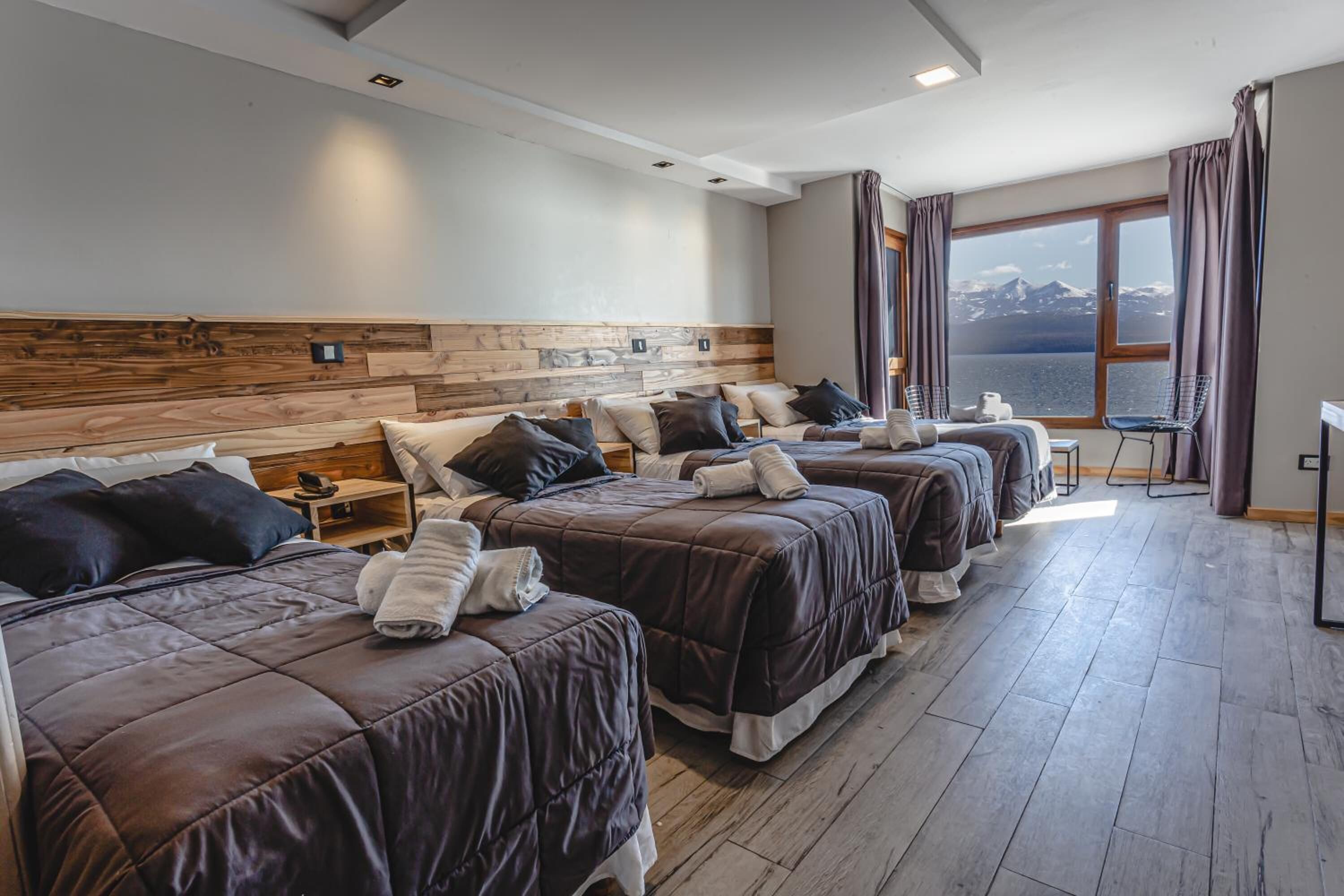 Habitación de hotel con cuatro camas dispuestas en fila, un ventanal con vista al mar y un cabecero de madera.