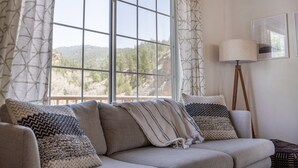 Un salon chaleureux avec une grande fenêtre, un canapé agrémenté de coussins à motifs, une lampe sur pied et un tableau encadré au mur.