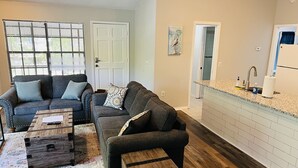 Ein Wohnzimmer mit zwei Sofas, einem Couchtisch und einem Teppich. Es gibt eine Küchenzeile mit Spüle und Arbeitsplatte.