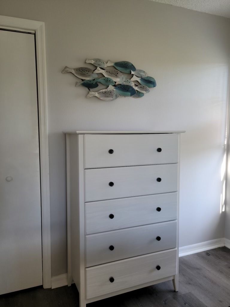 A white dresser with five drawers and black knobs, positioned next to a white door and a wall with a decorative fish art piece.
