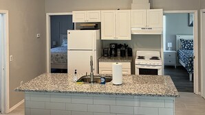 Eine moderne Küche mit weißen Schränken, einer Granitabdeckung und einer zentralen Kochinsel. Ein Kühlschrank, ein Herd und ein Spülbecken sind vorhanden. Die Küche ist durch Einbaustrahler gut beleuchtet.