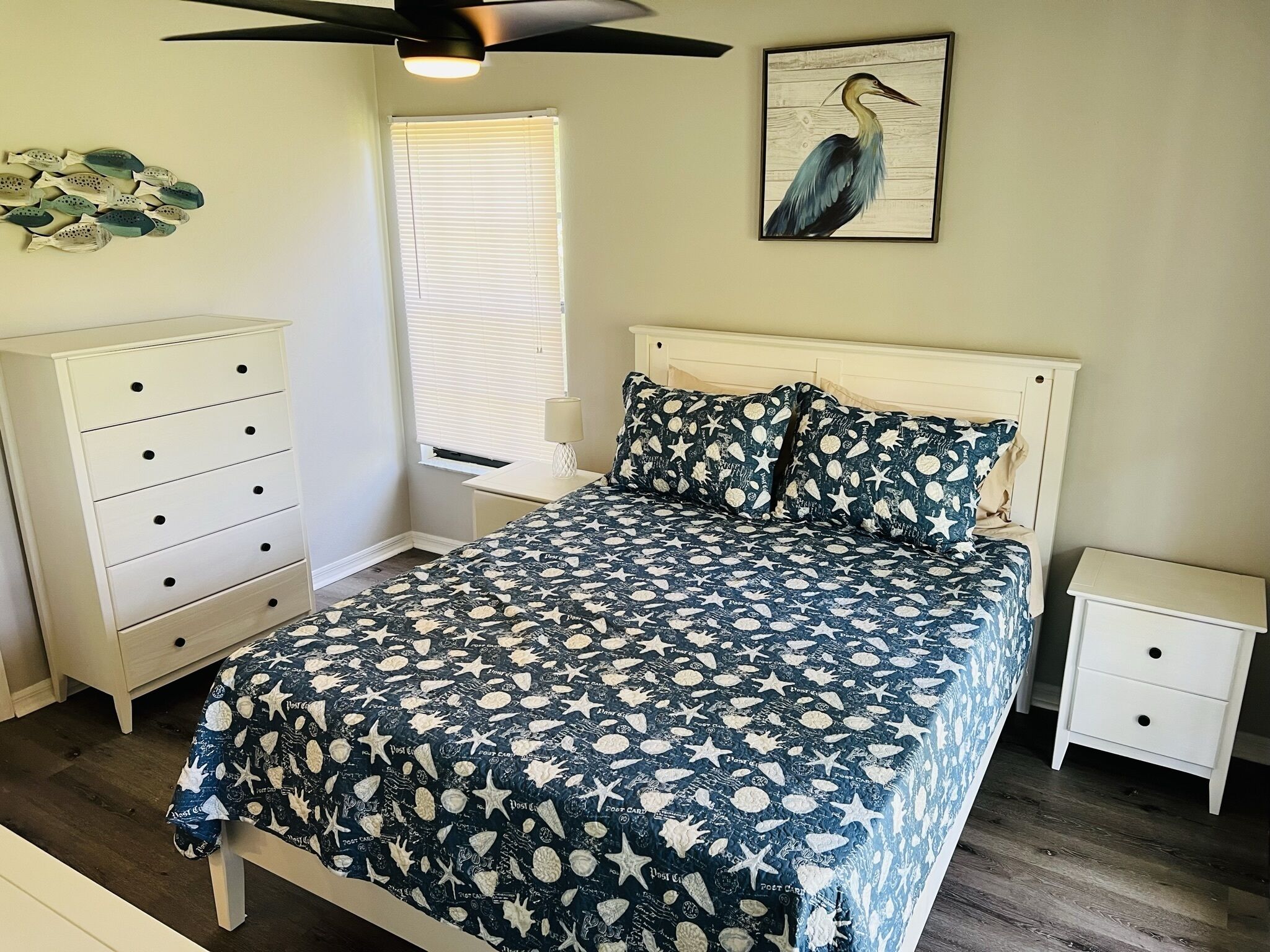 Ein Schlafzimmer mit einem Bett, einem Nachttisch, einer Kommode, einem Deckenventilator, einem Fenster mit Jalousien und einem Wandbild.