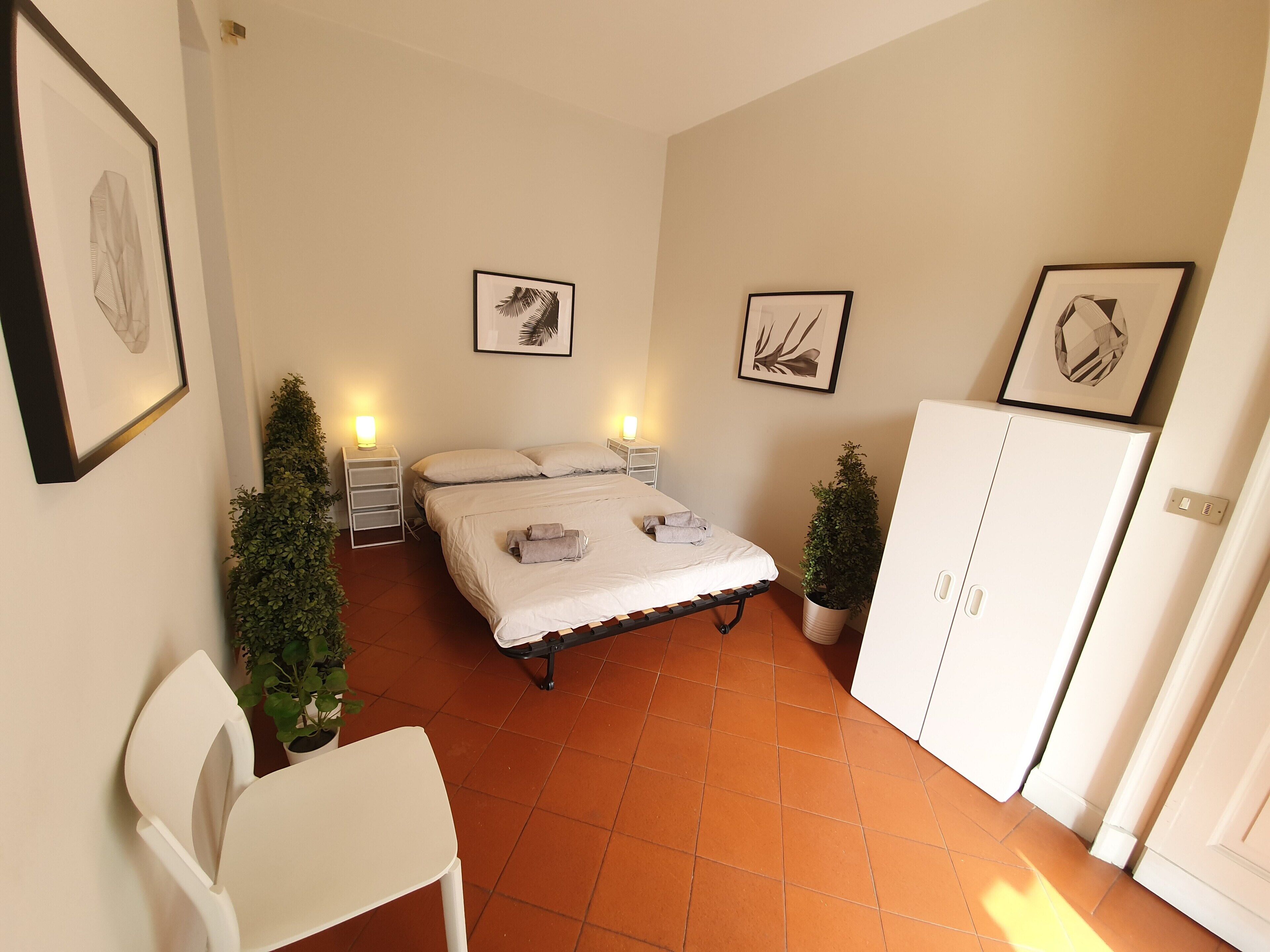 Un dormitorio con cama, mesitas de noche, una silla, armario y cuadros en las paredes.
