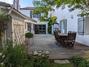 A courtyard with a wooden deck, a dining table with chairs, and a stone wall. - Saint Martin Intra Muros, Maison Calme Proche Commerces et Plages (Saint-Martin-de-Ré)