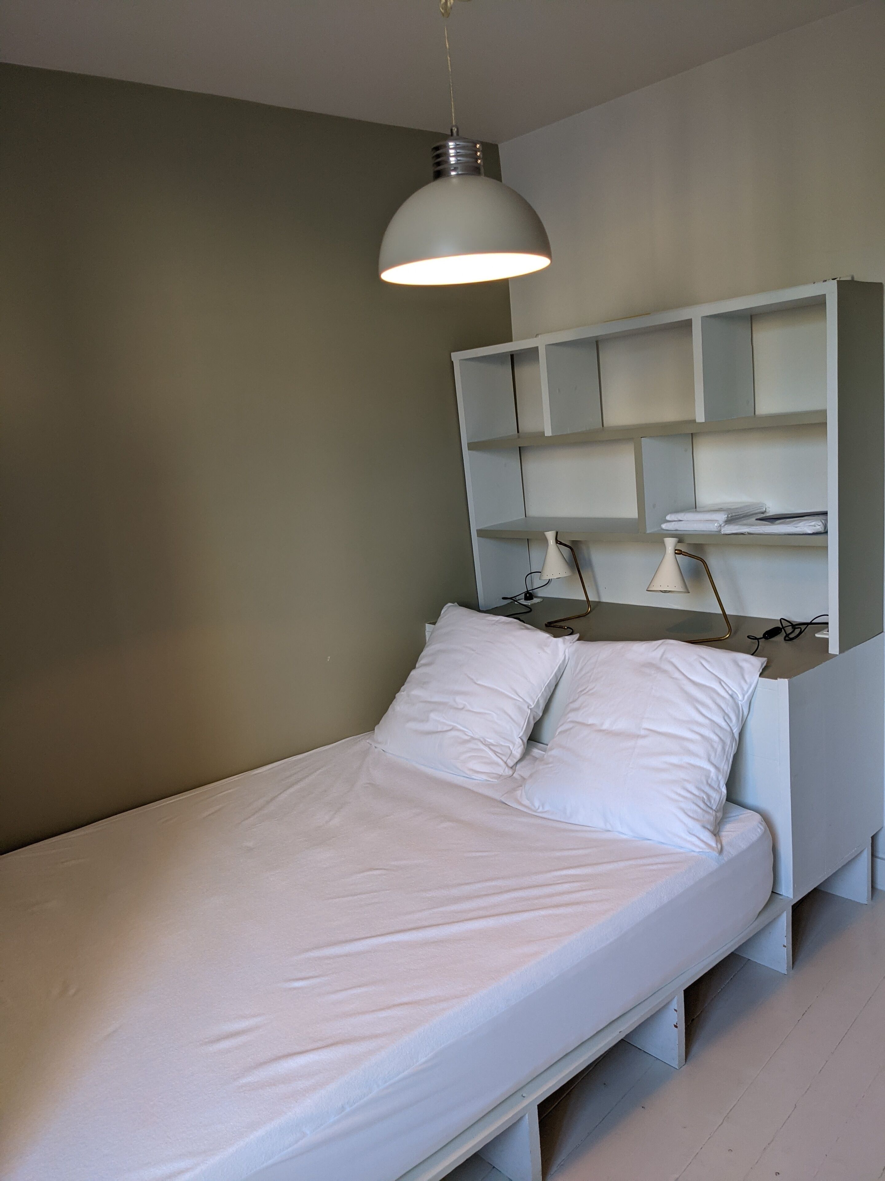 Un lit simple avec une literie et des oreillers blancs, une tête de lit blanche et une suspension lumineuse au-dessus.