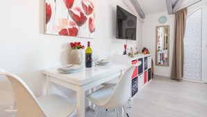 Un espace de salle à manger moderne avec une table blanche, des chaises et un grand tableau abstrait. Un téléviseur est fixé au mur, et un miroir est visible à l’arrière-plan.