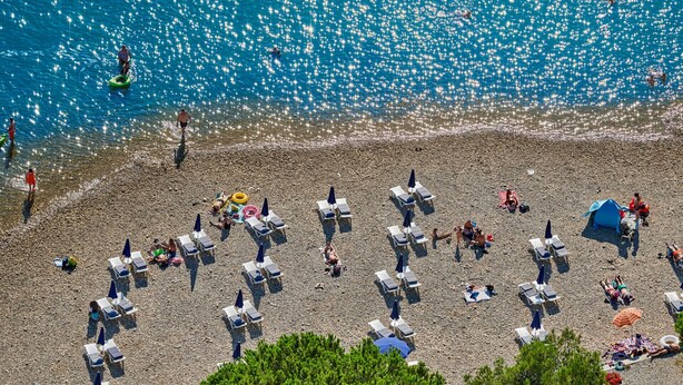 Une plage avec des chaises longues et des parasols, un rivage rocheux, et des gens qui se baignent et se promènent.