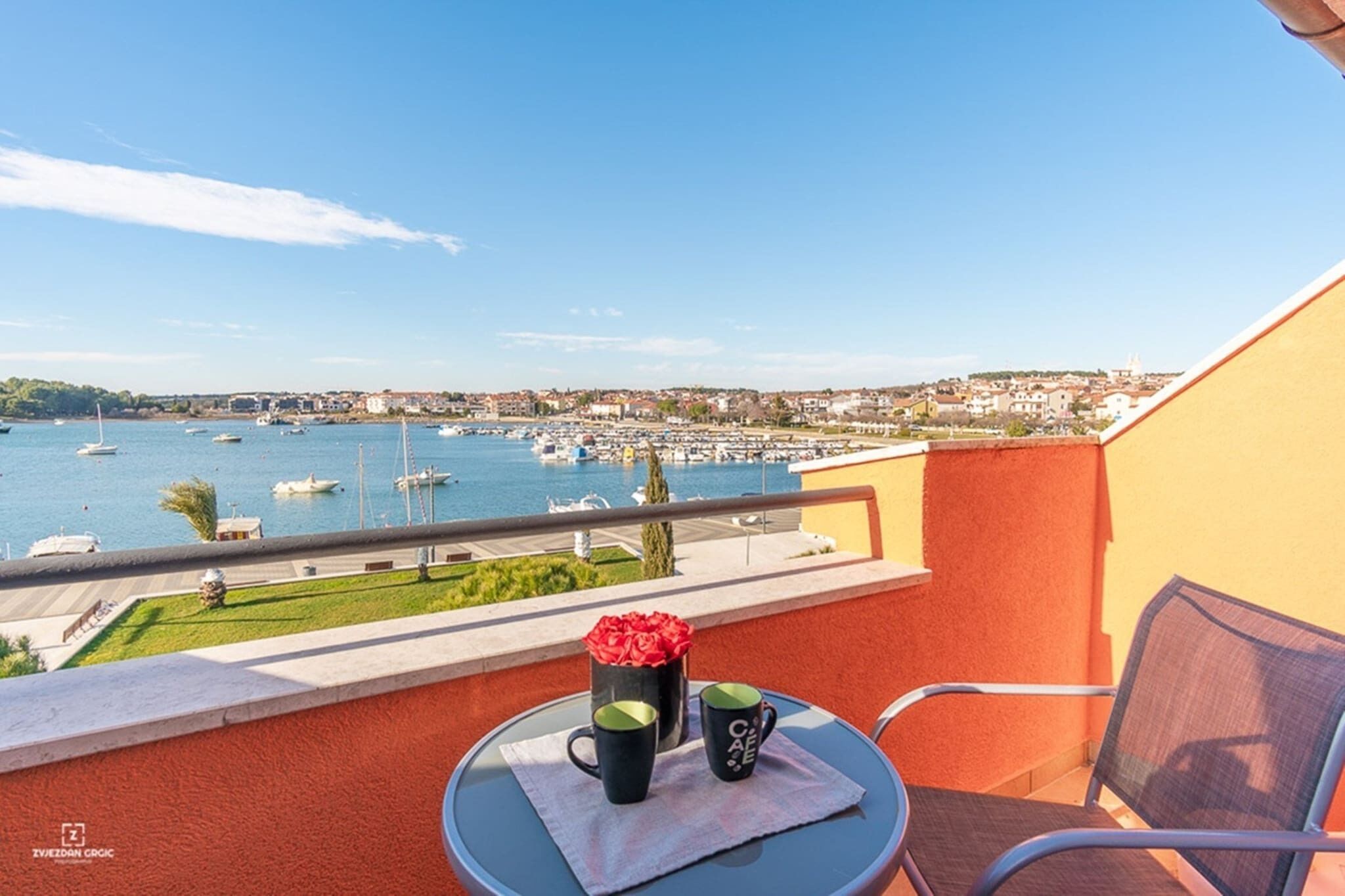 A balcony with a table, two mugs, and a vase of red flowers. The view includes a marina with boats and a distant town.