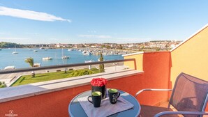 Un balcon avec une table, deux tasses et un vase rempli de fleurs rouges. La vue comprend un port de plaisance avec des bateaux et une ville située au loin.
