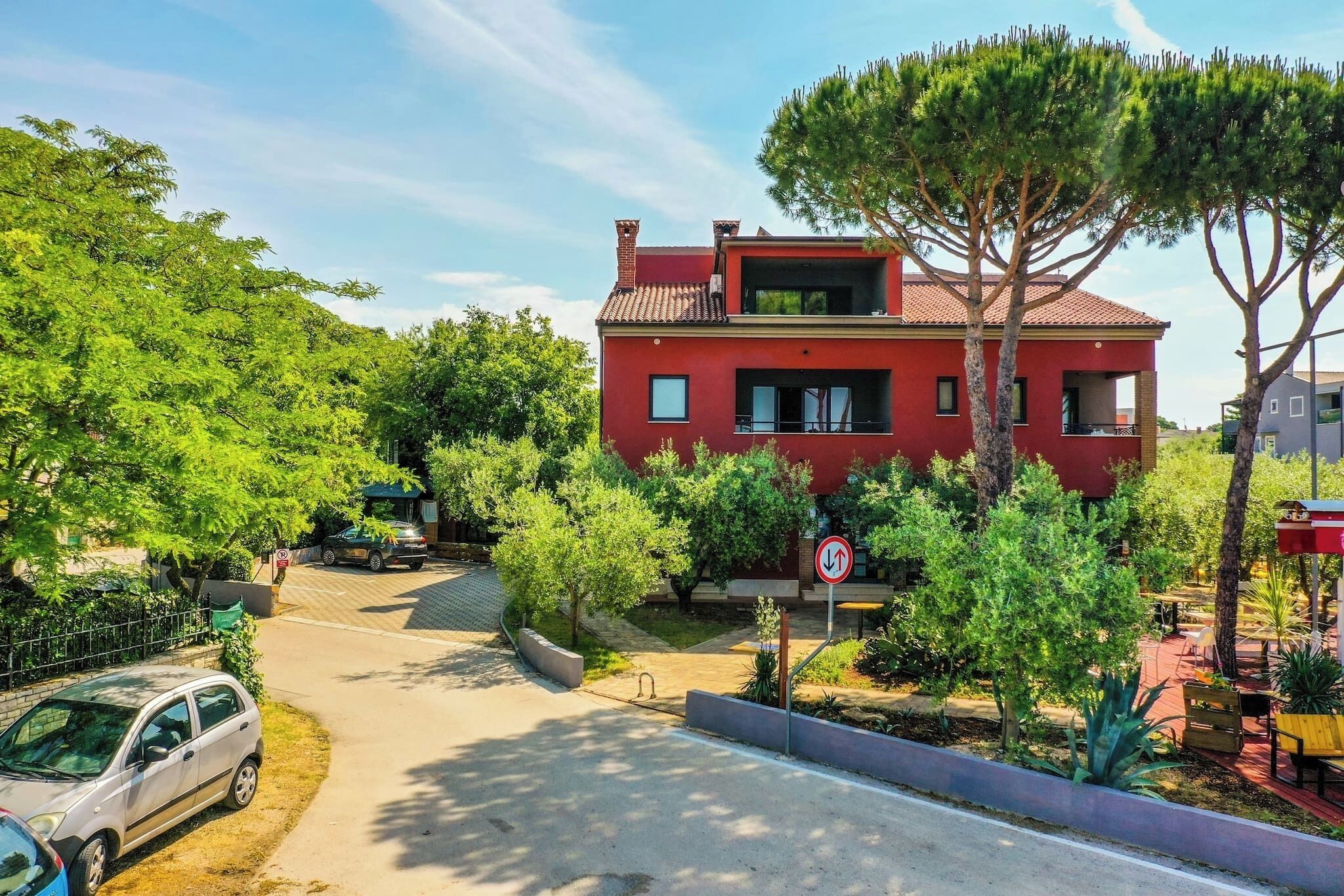 A red two-story house with a balcony, surrounded by greenery and a driveway.