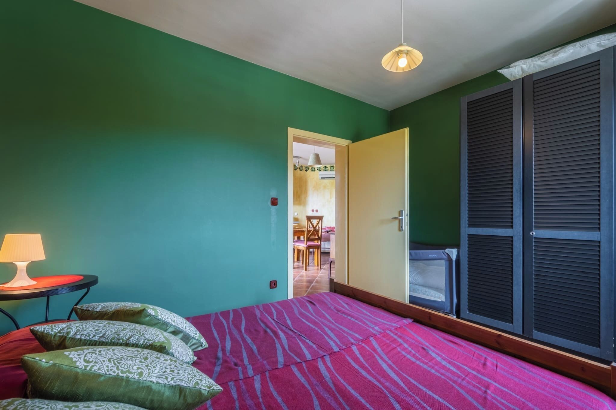 A room with a bed, a table with a lamp, and a door leading to another room.