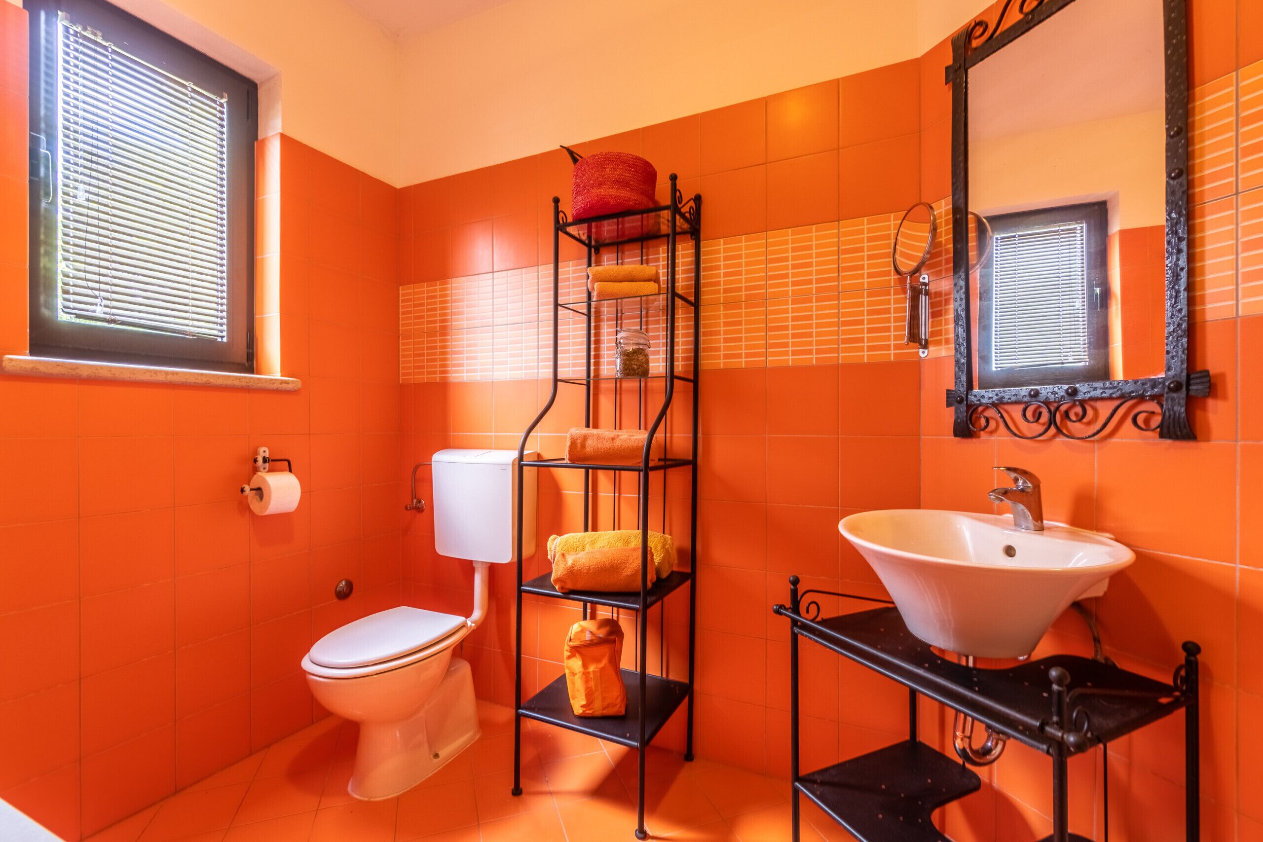 A bathroom with orange tiles, a white sink, a toilet, and a metal shelf unit with towels and toiletries.