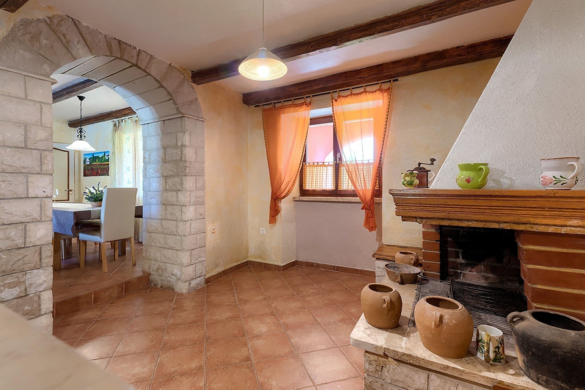 A rustic kitchen with a stone archway, a dining area with a table and chairs, and a brick fireplace with pottery.