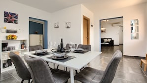 Un comedor moderno con una mesa blanca dispuesta para seis personas, rodeada de sillas grises. Se ve una cocina al fondo con una encimera negra y un refrigerador.