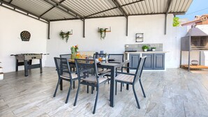 Un patio techado con una mesa de comedor, sillas, una mesa de fútbol de mesa, una encimera de cocina y plantas colgantes.