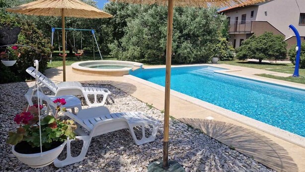 Une piscine entourée de chaises longues et d’une balançoire, le tout noyé dans la verdure, avec un bâtiment visible au loin.