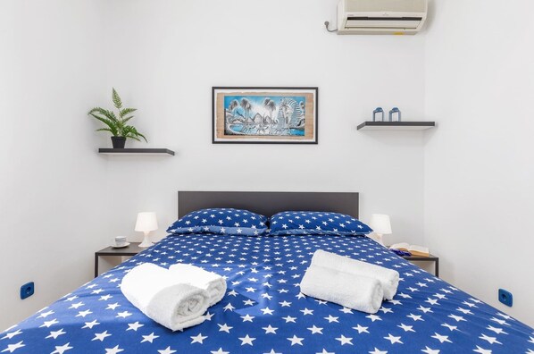 A hotel room with a bed featuring a blue and white star pattern, a framed painting on the wall, a small plant on a shelf, and a nightstand with a lamp and a book. - Sabi Relax Zone Apartment - Private Pool, Jacuzzi (Pula)