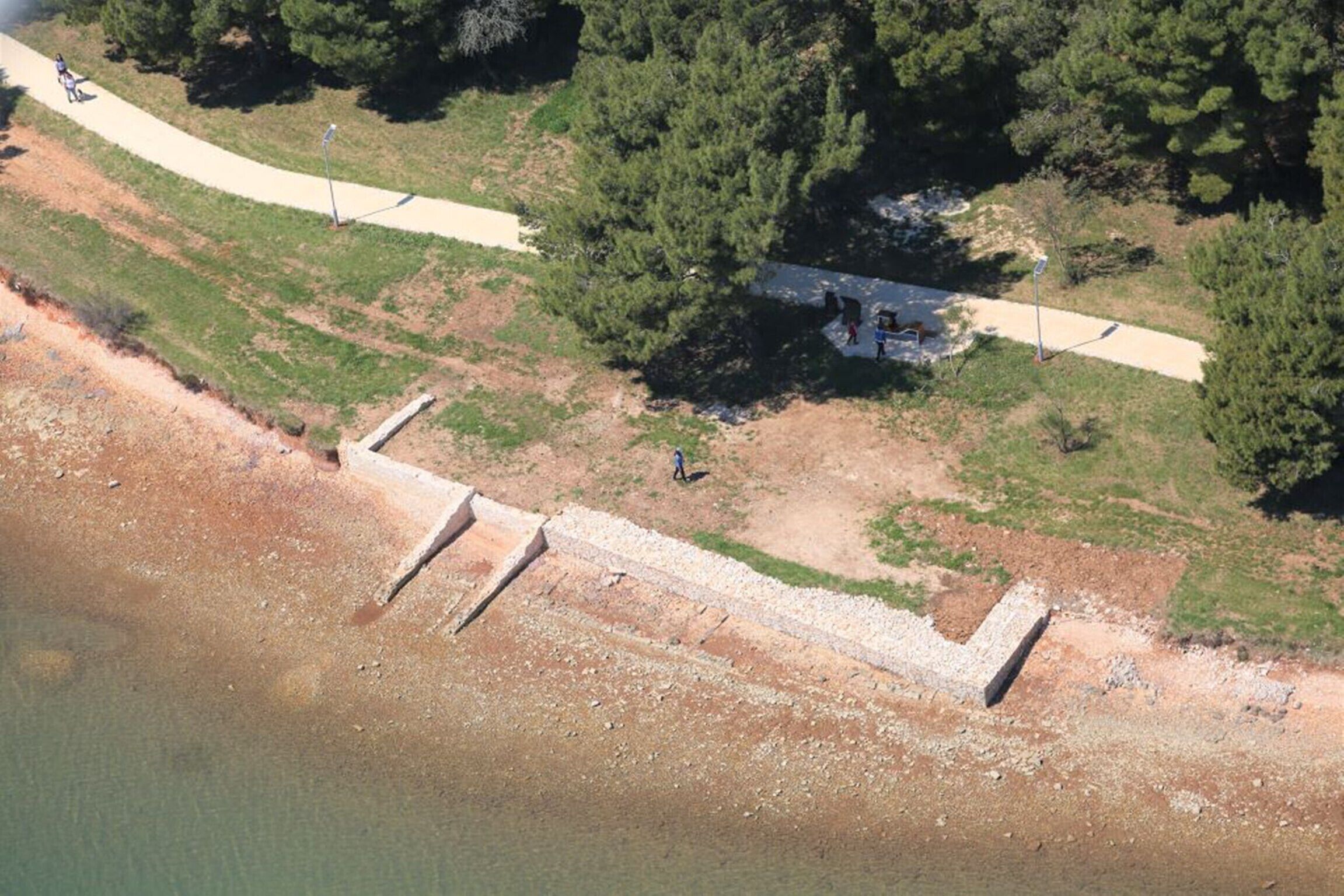An aerial view of a coastal area with a paved walkway, a small building, and a rocky shoreline.