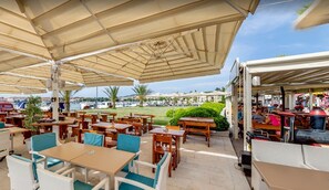 An outdoor cafe with wooden tables and chairs, covered by large umbrellas, overlooking a marina with boats and greenery.