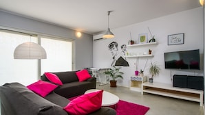 Un salon moderne avec un canapé gris, des coussins rouges, une table basse blanche, un tapis rose, un téléviseur et un mur orné d’une fresque en noir et blanc.