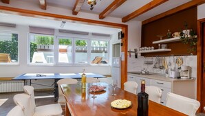 Un coin repas avec une table en bois, des chaises blanches, une vue sur le jardin grâce à de grandes fenêtres, et une cuisine équipée d’étagères et d’ustensiles.