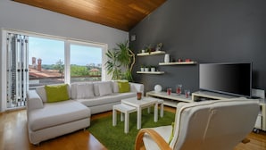 Un salon moderne avec un canapé blanc, des coussins verts, un téléviseur à écran plat et une grande fenêtre donnant sur la ville.