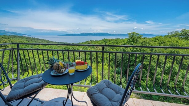 Un balcon avec une table dressée pour deux personnes, agrémentée d’un ananas, d’une pastèque et d’un verre de jus de fruits. Le balcon offre une vue sur un paysage verdoyant et luxuriant ainsi que sur une étendue d’eau.