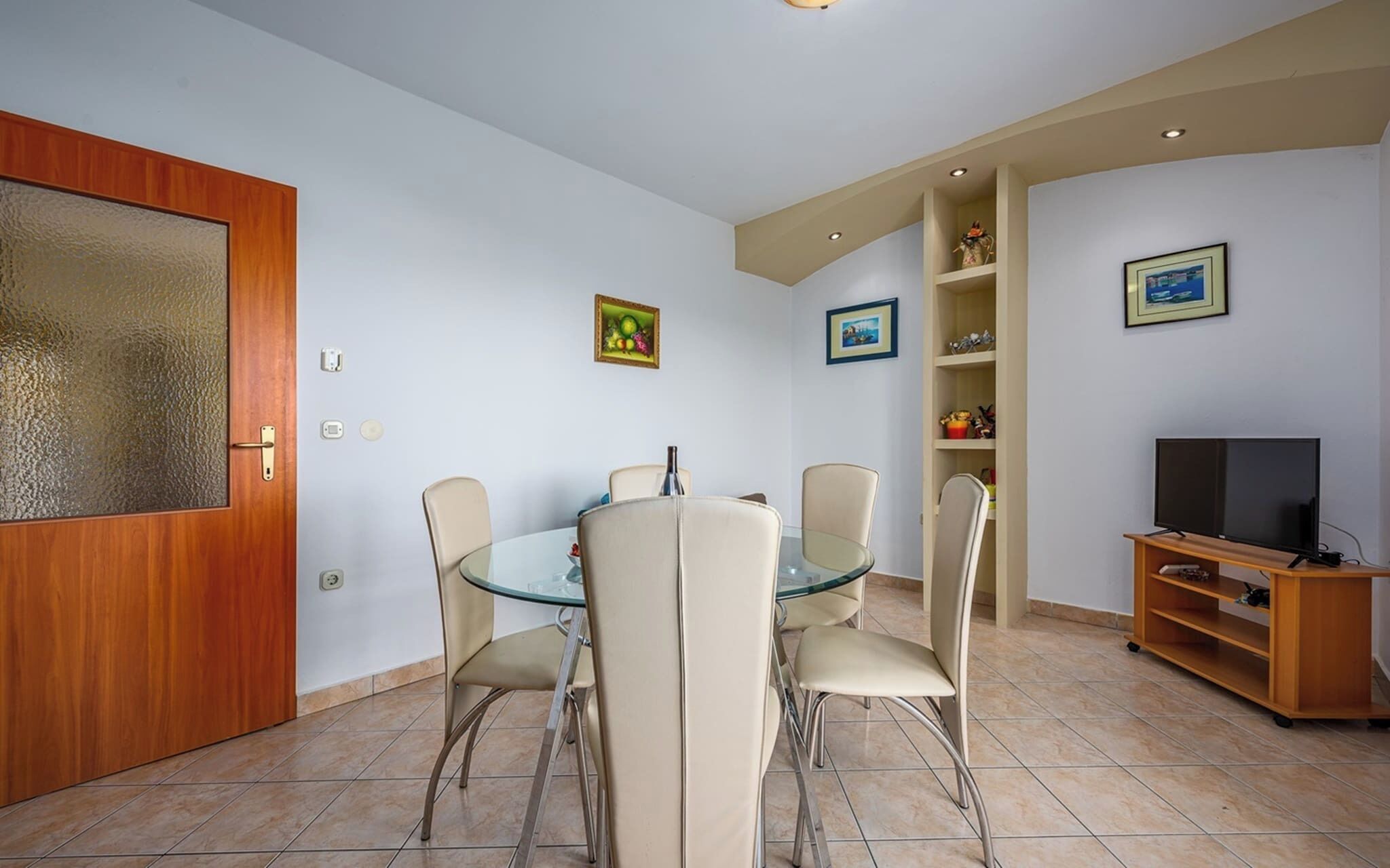 A dining area with a glass table and four chairs, a wooden door with a frosted glass panel, a television on a wooden stand, and a shelf with decorative items.