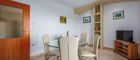A dining area with a glass table and four chairs, a wooden door with a frosted glass panel, a television on a wooden stand, and a shelf with decorative items.