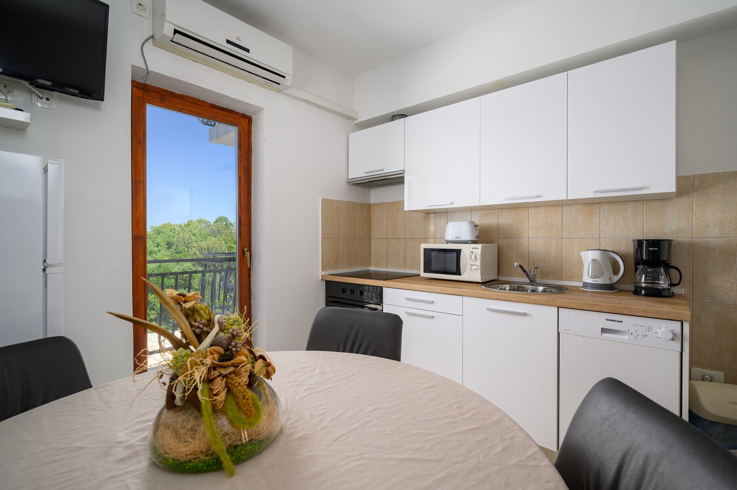 A modern kitchen with white cabinets, a microwave, a coffee maker, and a dishwasher. There is a dining table with a vase of dried flowers.