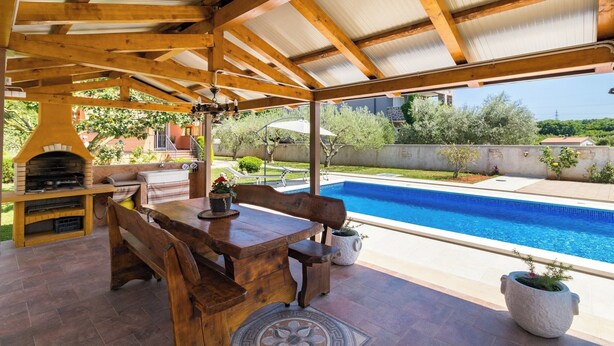 Een terras buiten met een houten tafel en banken, een barbecue en een zwembad.