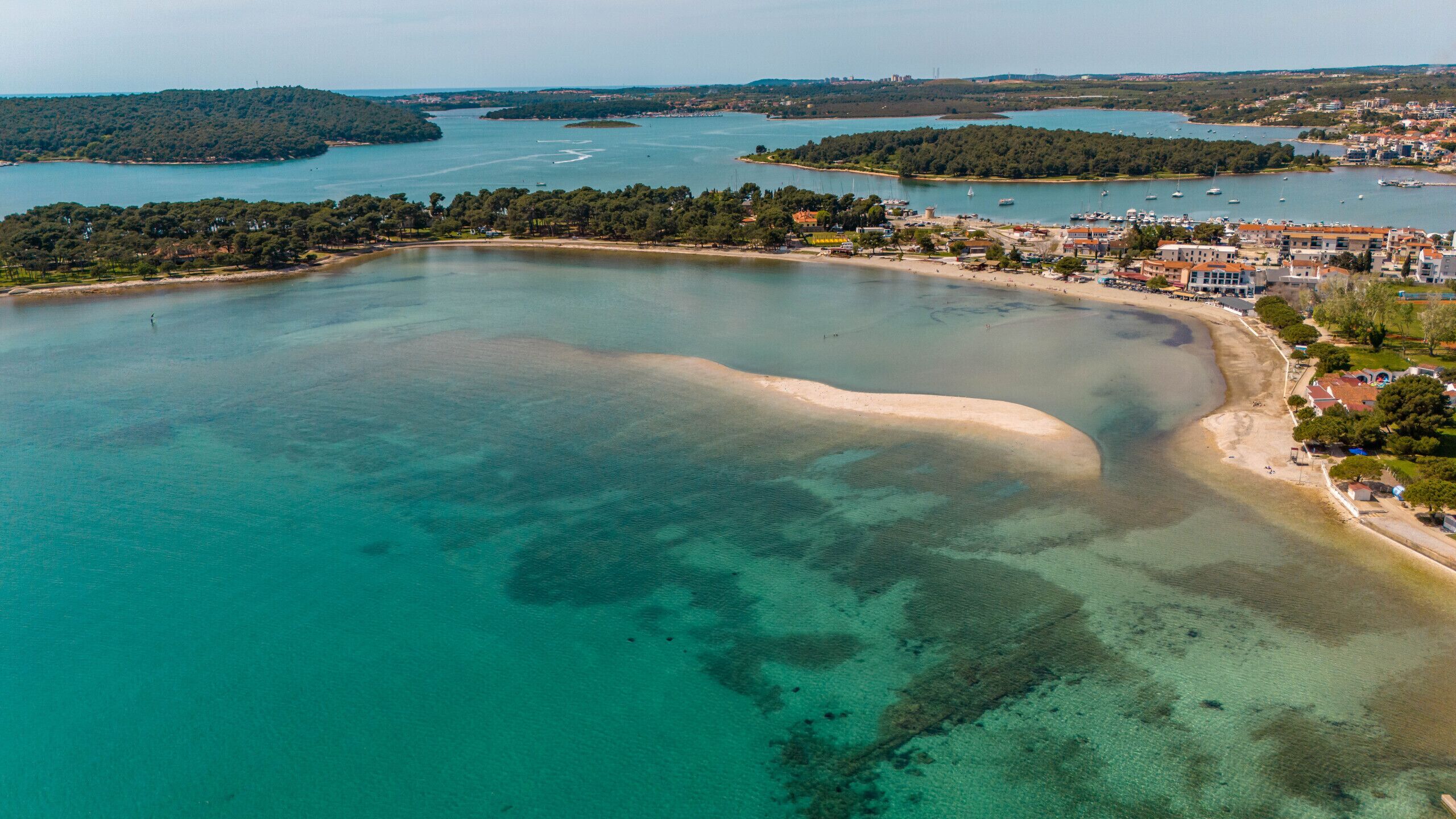 Vue aérienne d’une zone côtière avec des eaux cristallines, une plage de sable et une ville dont les bâtiments s’étendent le long du littoral.