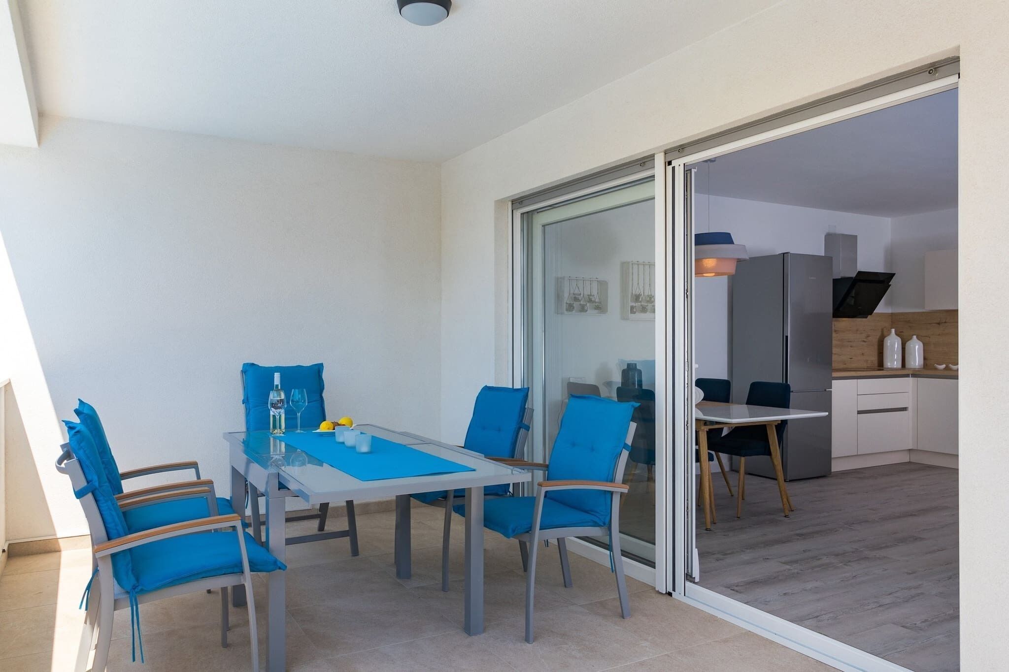 Een eetruimte met een glazen tafel en blauwe stoelen, een keuken met witte kastjes en een schuifdeur van glas naar een andere ruimte.
