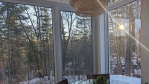 Une table en verre, des chaises et une plante, avec vue sur une forêt enneigée à travers de grandes fenêtres.