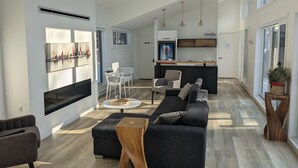Un salon moderne comprenant un canapé, un pouf et une table basse, un coin repas avec des chaises, une cuisine avec un bar et de grandes fenêtres qui laissent entrer la lumière naturelle.