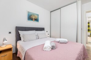 En pænt redt seng med et pink sengetæppe, to sammenrullede håndklæder og en grå sengegavl.