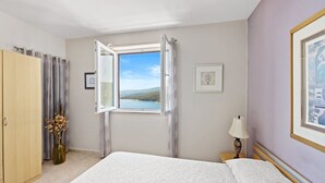 Ein Schlafzimmer mit Bett, einem Fenster mit Vorhängen, einem Nachttisch mit Lampe und einer Vase mit getrockneten Blumen.