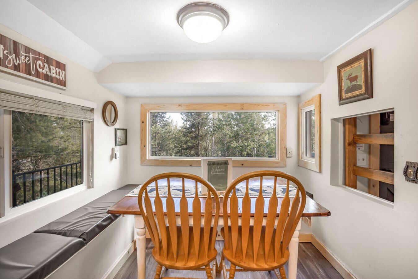 A cabin interior with a wooden bench, two wooden chairs, a window with a view of trees, and a sign reading "sweet cabin".