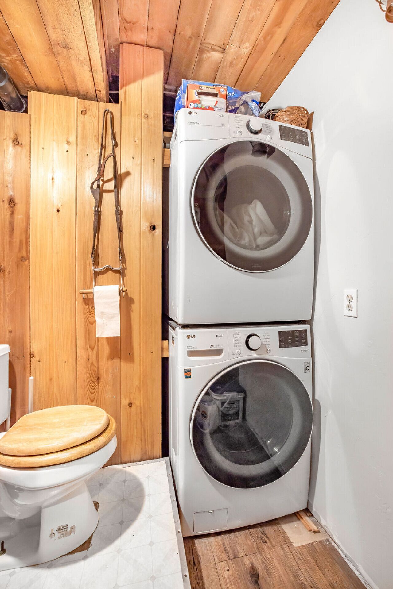 Een kleine wasruimte met houten plafond en wanden, een wasmachine, een droger en een toilet met houten zitje.
