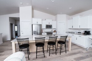 A modern kitchen with white cabinets, a stainless steel refrigerator, a microwave, and a granite countertop. There are four black chairs at the island.