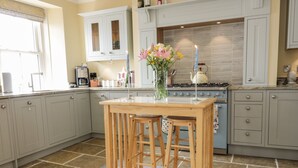 Una cocina con una mesa de madera, dos taburetes de bar, un jarrón con flores y una vela encendida.