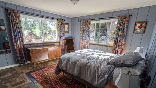 A bedroom with a bed, a dresser, a chair, a window with curtains, and a lamp. - Sleeping Wolf Guest House (Oakhurst)